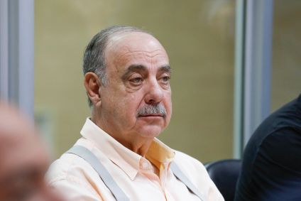 Fuad Noman é um homem branco, de bigode e cabelos brancos. Ele tem olhos claros e na imagem usa uma camisa social laranja claro com um suspensório cinza.