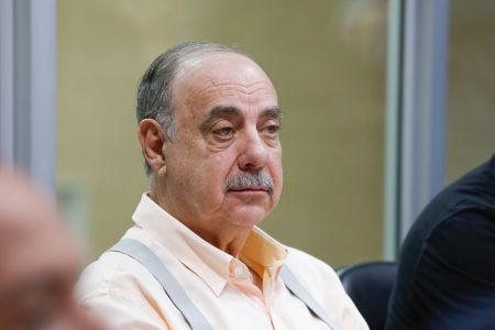 Fuad Noman é um homem branco, de bigode e cabelos brancos. Ele tem olhos claros e na imagem usa uma camisa social laranja claro com um suspensório cinza.