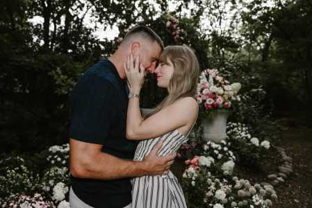 Uma foto de close de Taylor Swift e Travis Kelce em um jardim. Eles estão de frente um para o outro, com as testas encostadas e os olhos fechados, parecendo emocionados. Taylor usa um vestido listrado e está com uma das mãos no rosto de Travis. Travis, com um suéter azul marinho, a abraça pela cintura. O fundo desfocado mostra um arranjo floral grande em um vaso de pedra e folhagens.