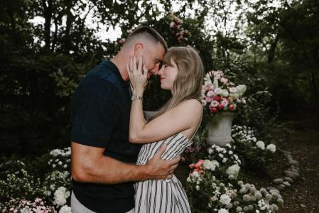 Uma foto de close de Taylor Swift e Travis Kelce em um jardim. Eles estão de frente um para o outro, com as testas encostadas e os olhos fechados, parecendo emocionados. Taylor usa um vestido listrado e está com uma das mãos no rosto de Travis. Travis, com um suéter azul marinho, a abraça pela cintura. O fundo desfocado mostra um arranjo floral grande em um vaso de pedra e folhagens.