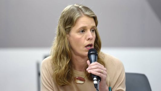 Uma mulher branca, de cabelo loiro, segura um microfone. Ela usa uma camisa bege e aliança dourada no dedo anelar.