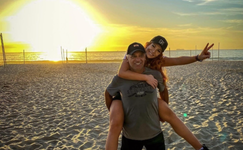 Na imagem, um homem de cabelos brancos, boné preto, bermuda preta e camisa cinza carrega nas costas uma mulher branca, de cabelos ruivos e boné vermelho. Ela faz o sinal de "paz e amor". Eles estão em uma praia com o por do sol atrás.