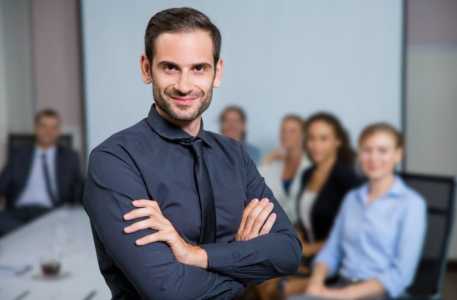 Homem sorrindo com terno sentado em uma mesa com colegas atrás