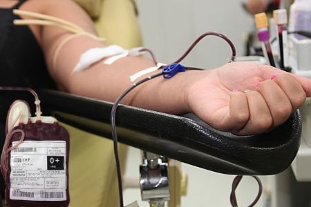 Pessoa doando sangue. Na imagem, é possível ver um braço de uma mulher com as unhas feitas. Há um dispositivo de retirada de sangue para doação em seu braço e uma bolsa de sangue O positivo  próximo