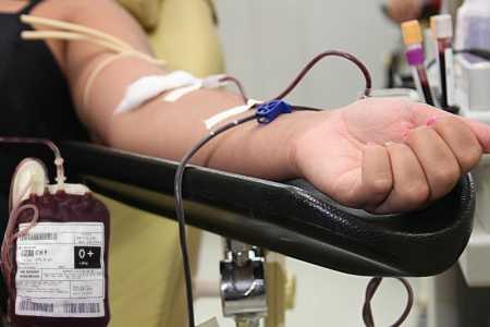 Pessoa doando sangue. Na imagem, é possível ver um braço de uma mulher com as unhas feitas. Há um dispositivo de retirada de sangue para doação em seu braço e uma bolsa de sangue O positivo  próximo