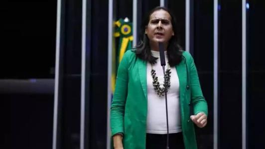 Uma mulher de cabelos pretos usa uma camisa branca, um blazer verde e um colar prateado.
