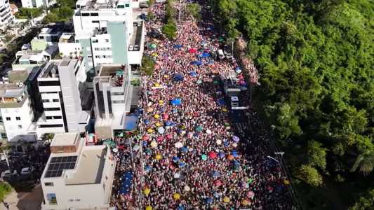 Vista aérea da Avenida Altamiro Soares, no bairro Castelo, da região da Pampulha de Belo Horizonte. Do lado direito, uma região de floresta. No centro da imagem, na Avenida, milhares de pessoas ocupam a rua durante o Carnaval de 2024. É possível identificar o trio elétrico do grupo Baianeiros no canto direito e guarda-sóis de várias cores. Do lado esquerdo, os prédios da Avenida.