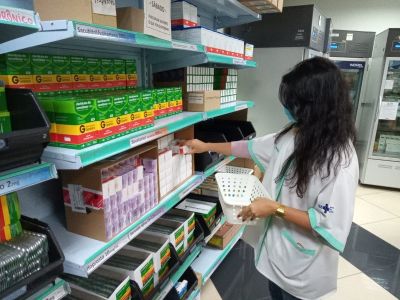 Uma mulher, de pele branca e cabelos longos, vestida com um colete do SUS, pega uma caixa de remédio de uma prateleira de uma farmácia