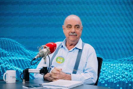 Fuad Noman é um homem branco, de bigode e cabelos brancos. Ele tem olhos claros e na imagem usa uma camisa social laranja claro com um suspensório cinza.