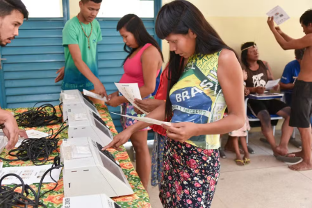 Na imagem, uma mulher indígena de cabelos longos, lisos e pretos segura um papel enquanto vota em uma urna eletrônica. Ela usa uma camisa sem mangas estampada com a bandeira do Brasil e uma saia preta com rosas cor de cosa e folhas verdes.