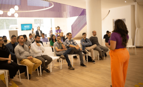 Mulher de cabelos castanhos, com blusa roxa e calça laranja, palestra para um grupo de homens, sentados em cadeiras brancas na sede do Órbi Conecta