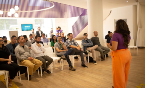 Mulher de cabelos castanhos, com blusa roxa e calça laranja, palestra para um grupo de homens, sentados em cadeiras brancas na sede do Órbi Conecta