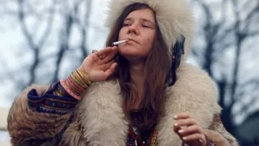 Janis Joplin aprecia um cigarro vestida com um casaco de pele e inúmeros badulaques em seus braços
