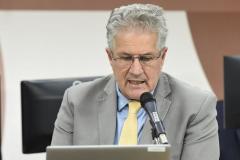 João Leite é um homem branco de cabelos brancos. Na imagem, ele usa óculos de grau, fala em frente a um microfone enquanto olha para um notebook cinza e usa terno cinza com gravata amarela.