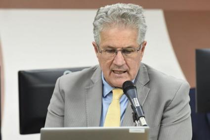 João Leite é um homem branco de cabelos brancos. Na imagem, ele usa óculos de grau, fala em frente a um microfone enquanto olha para um notebook cinza e usa terno cinza com gravata amarela.