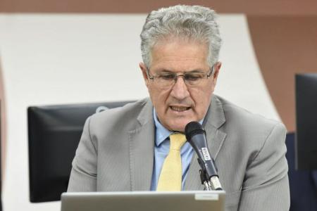 João Leite é um homem branco de cabelos brancos. Na imagem, ele usa óculos de grau, fala em frente a um microfone enquanto olha para um notebook cinza e usa terno cinza com gravata amarela.
