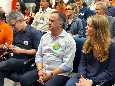 Mauro Tramonte, no canto esquerdo. Ele é um homem branco, de cabelo grisalho. Na imagem, usa uma camisa social azul clara com um adesivo verde, amarelo e azul. Ao lado, Luísa Barreto. Ela é uma mulher branca, loira e usa uma camisa social azul escura. Ao fundo, várias pessoas usando camisas laranjas, e uma usando uma camisa amarela segura uma câmera.