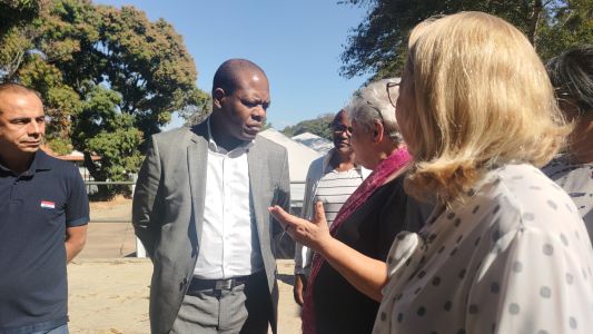 Ministro Almeida. Ele é um homem preto, careca, e na imagem conversa com duas mulheres com as mãos para trás. Almeida usa terno cinza e blusa social branca.