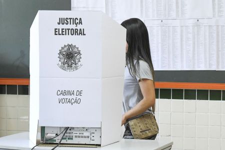 Mulher branca de cabelo escuro, camisa cinza e bolsa marrom indo para a cabine de votação eleitoral.