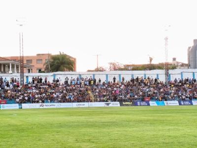 Foto mostra torcedores sentados em uma arquibancada em frete a um campo de futebol