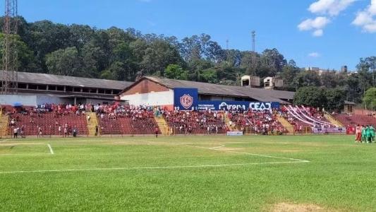 Imagem mostra o gramado do campo de futebol do Estádio Israel Pinheiro. Ao fundo estão pessoas em uma arquibancada vermelha e amarela, observando os jogadores reunidos em campo