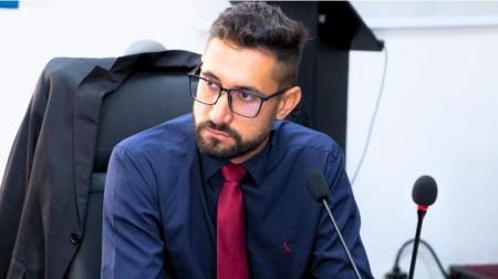 Paulo Bigodinho é um homem branco, de barba e cabelos castanho escuro, usa óculos de grau com alça preta, e na imagem ele sorri enquanto usa uma camisa social branca clara.