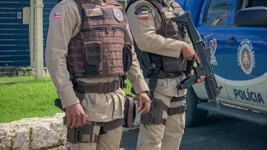 Dois policiais militares da Bahia aparecem de pé. Um deles está com uma metralhadora em mãos