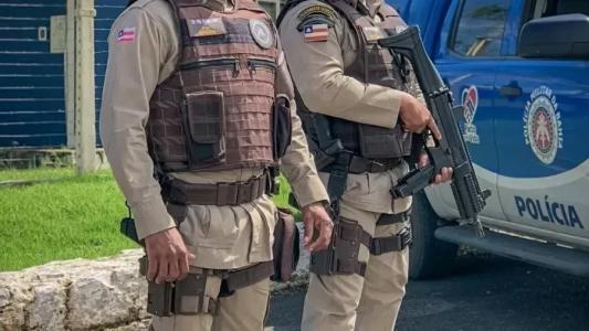 Dois policiais militares da Bahia aparecem de pé. Um deles está com uma metralhadora em mãos