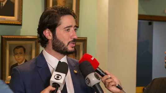 Gabriel Azevedo é um homem branco, de cabelo preto e barba preta. Ele usa um terno azul escuro na imagem enquanto fala no microfone da Itatiaia.
