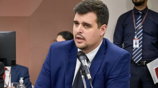 Homem branco com cabelo e barba de cor castanho escuro com terno azul marinho.