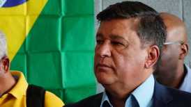 Homem branco, de cabelo cinza, usando roupas sociais. Bandeira do Brasil ao fundo.