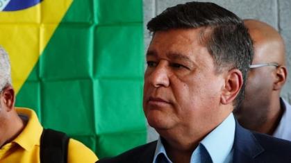 Homem branco, de cabelo cinza, usando roupas sociais. Bandeira do Brasil ao fundo.