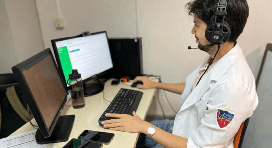 A imagem mostra um médico (um homem branco, de cabelo liso, na cor preta, e barba) sentado em frente à um computador, com um fone de ouvido na cabeça
