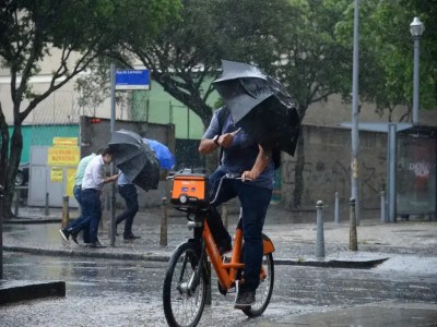 Chove forte em rua do Rio de Janeiro. Na imagem, um homem em uma bicicleta laranja, no primeiro plano, se equilibra e tenta se proteger com um guarda-chuva. No fundo, três homens andam na chuva com guardas-chuva