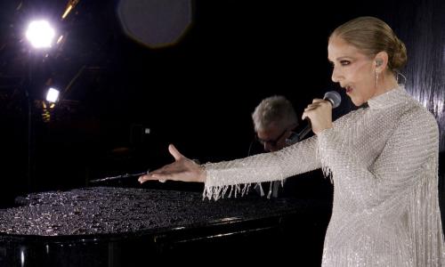 Celine Dion de vestido branco brilhante se apresentando nas Olimpíadas de Paris de 2024. No fundo, um homem tocando um piano de cauda preto.