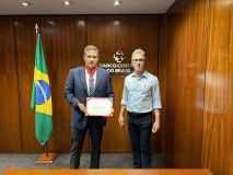 Na imagem, dois homens brancos estão de pé, ao lado da bandeira do Brasil, com uma parede de madeira ao fundo. No lado esquerdo, Roberto Campos Neto, presidente do Banco Central, usa terno, usa a medalha e segura um certificado. Do lado esquerdo, Romeu Zema, governador de Minas Gerais.