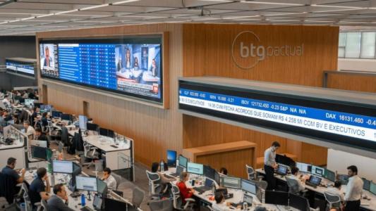Sede do BTG Pactual, com televisões mostrando números e operações comerciais. No escritório, na parte superior, há detalhes de madeira. Já na parte inferior da foto, pessoas trabalham em mesas
