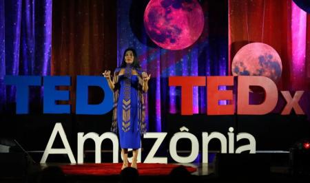Palestrante com vestes indígenas azuis no palco do TEDxAmazônia. A iluminação é azul, na parte esquerda, e vermelha, na parte direita