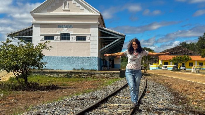 Imagem mostra a curadora da exposição, Anna Veloso, sob os trilhos da ferrovia de Bocaiúva. Ao fundo está o Centro de Memória Casa Figueiredo Souza 