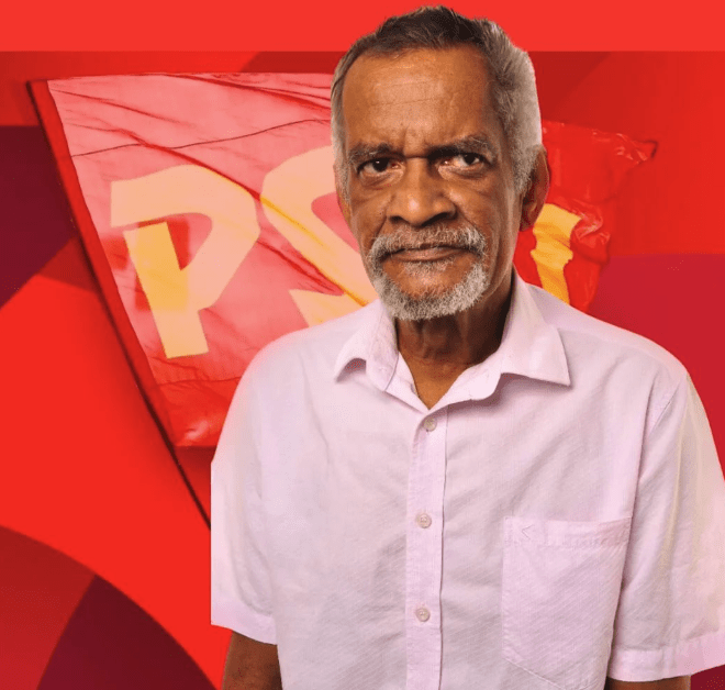 Gilberto Cunha é um homem preto, de cabelos e barba branca. Na imagem, ele usa uma camisa social de manga curta de cor rosa claro. Ao fundo, uma bandeira vermelha.