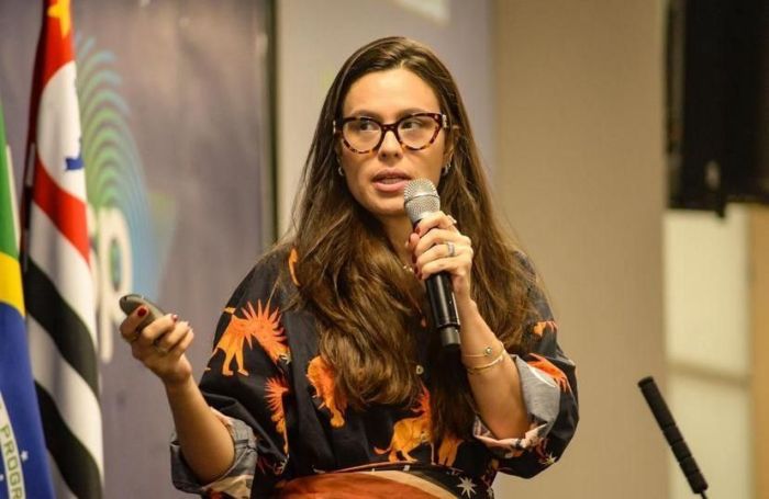 Na imagem, uma mulher branca de cabelos cumpridos castanhos e de óculos, segura um microfone enquanto usa uma camisa de botão preta estampada com leões.