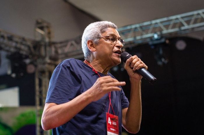 Cyro Garcia é um homem negro, de cabelos brancos, usa óculos. Na imagem, ele segura um microfone e usa camisa azul escura com um cordão de identificação vermelho.