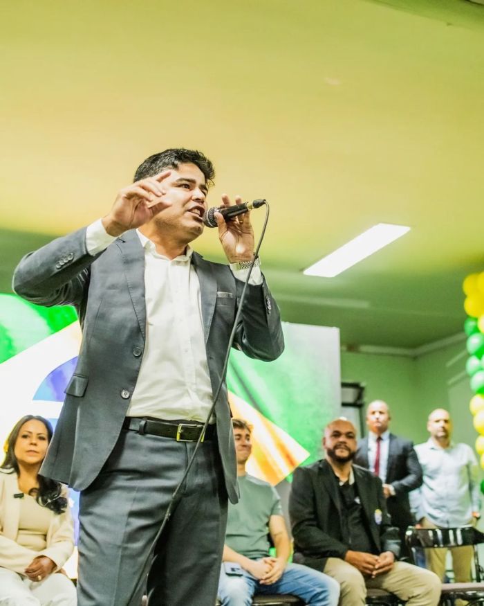 Dadson é um homem branco, usa terno cinza e blusa social branca enquanto fala em um microfone.