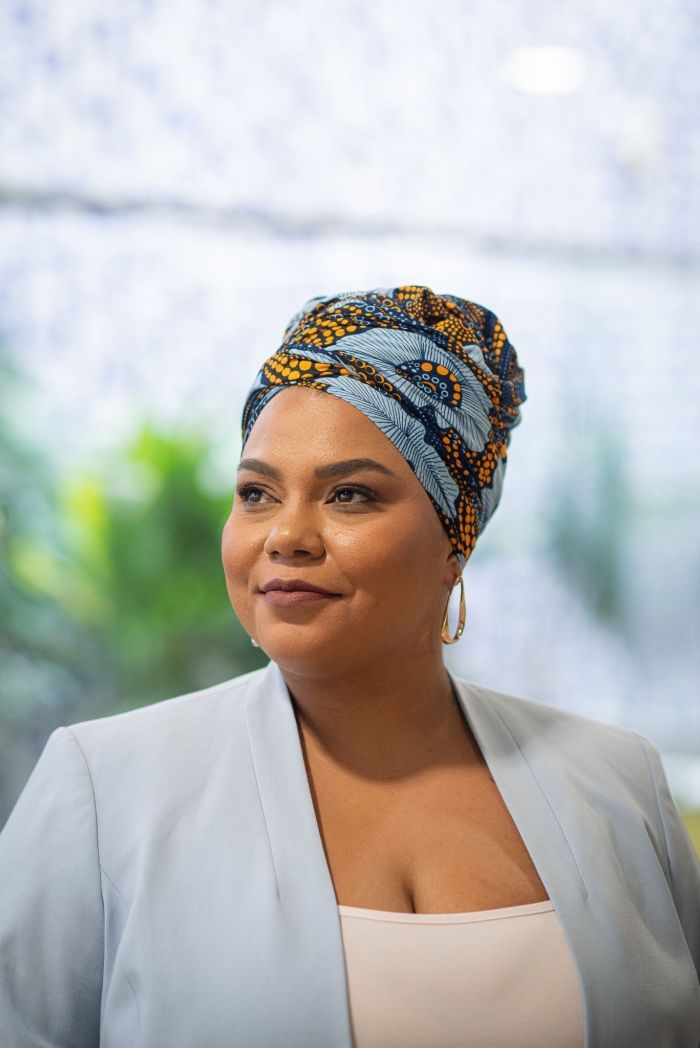 Dandara é uma mulher preta. Na imagem, ela usa um tubante azul-claro com detalhes amarelos. Ela usa uma argola dourada, um terno azul bebê e uma camiseta branca.