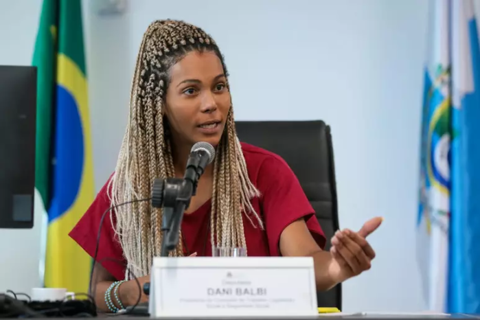 Dani Balbi é uma mulher preta, de cabelos trançados e loiros. Na imagem, ela fala em um microfone usando pulseiras e uma blusa vermelha. 