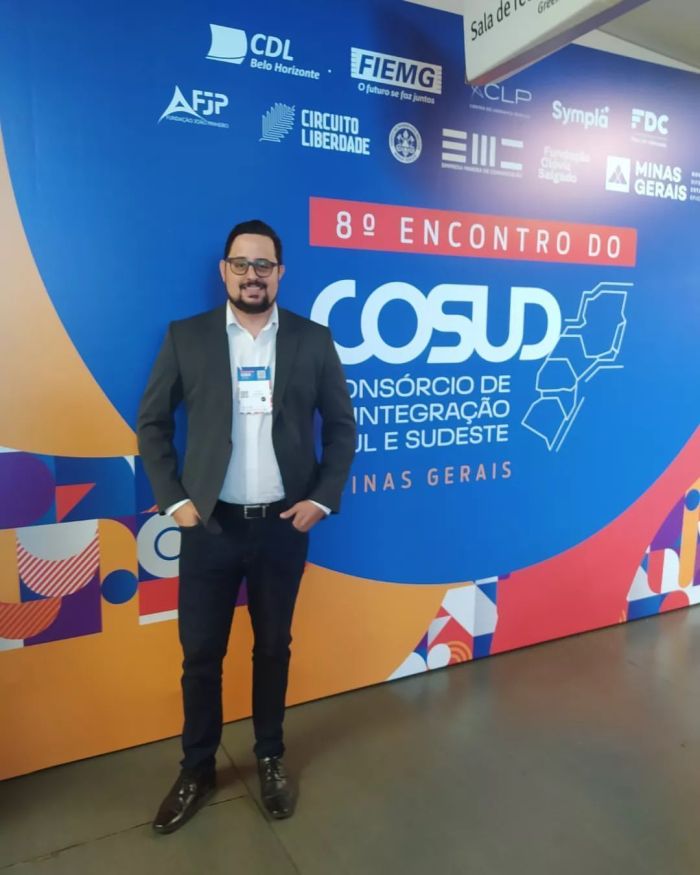 Diogo Fernandes é um homem branco, de óculos de grau, cabelos e barba preta. Ele usa um terno cinza, camisa social branca e calça social preta.