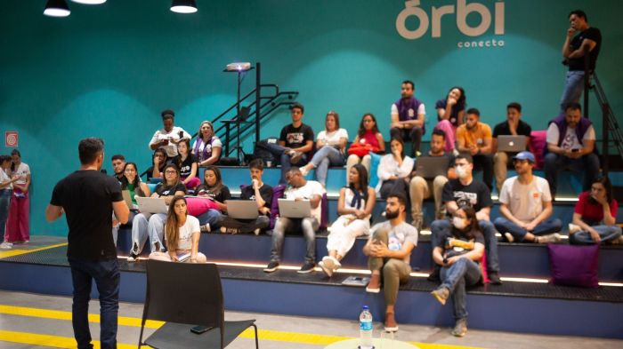 Auditório do Órbi Conecta, localizado em Belo Horizonte, com pessoas sentadas em uma arquibancada, enquanto um homem de blusa preta palestra