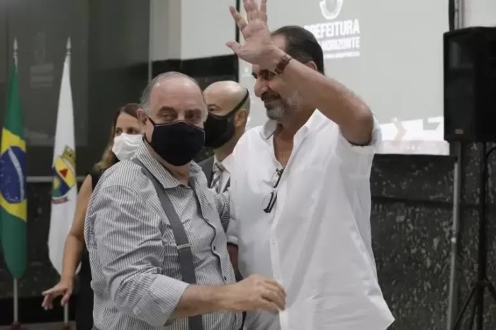 Na imagem, Fuad Noman está no canto esquerdo, abraçando Alexandre Kalil. Ele é um homem branco, de cabelos brancos, usa máscara preto que cobre o nariz e a boca, camisa listrada com suspensório. Alexandre Kalil usa uma camisa branca, óculos de grau pendurado na camisa, relógio e acena. 