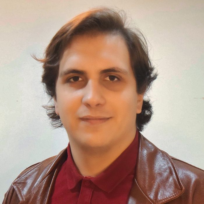Henrique Simonard é um homem branco de cabelo marrom até a altura do ombro. Na imagem, ele usa uma jaqueta de couro marrom com uma blusa de botão vermelha.
