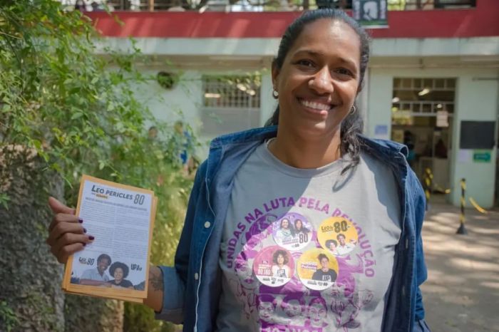 Indira Xavier é uma mulher preta de cabelos escuros e lisos. Ela usa uma camisa cinza com escritos e desenhos roxos, jaqueta jeans e segura um panfleto. Está com o cabelo preso em um rabo de cavalo.